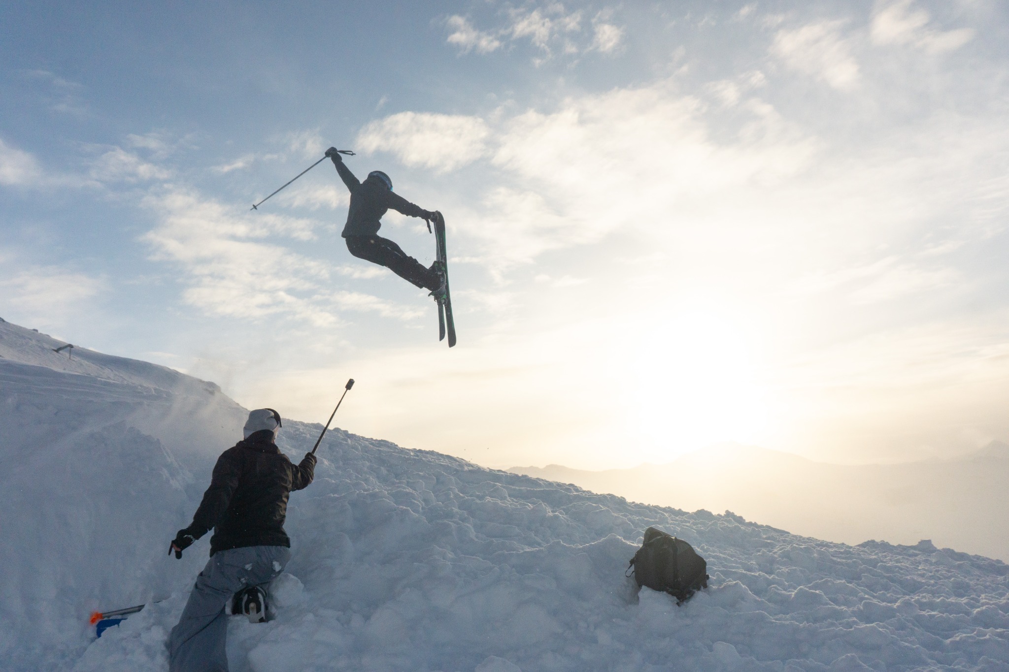 A skier airborne in golden mountain light, with a friend filming the jump below